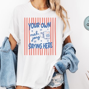 May include: A white short-sleeve t-shirt featuring a graphic with vertical coral-colored stripes. The text reads YOUR OWN mah jong SAYING HERE in blue, accompanied by a small illustration of a mahjong tile and a box labeled JOKER.