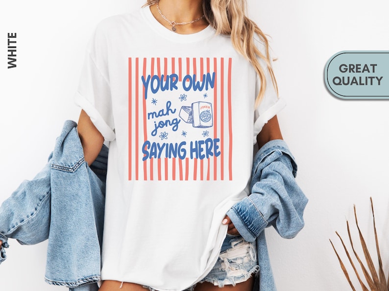 May include: A white short-sleeve t-shirt featuring a graphic with vertical coral-colored stripes. The text reads YOUR OWN mah jong SAYING HERE in blue, accompanied by a small illustration of a mahjong tile and a box labeled JOKER.
