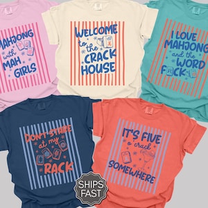 May include: Five short-sleeved crew neck t-shirts in pink, cream, teal, navy, and coral. Each shirt features a central graphic with vertical stripes and text related to mahjong, including phrases like Mahjong with Mah Girls and It's Five o'crack Somewhere.