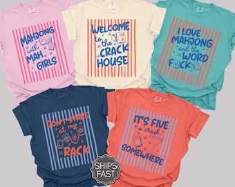 May include: Five short-sleeved crew neck t-shirts in pink, cream, teal, navy, and coral. Each shirt features a central graphic with vertical stripes and text related to mahjong, including phrases like Mahjong with Mah Girls and It's Five o'crack Somewhere.