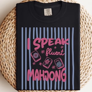 May include: A black t-shirt featuring a graphic with vertical light blue stripes and the pink text "I SPEAK fluent MAHJONG". The design includes illustrations of three mahjong tiles, including one labeled "JOKER", and small pink flower accents.