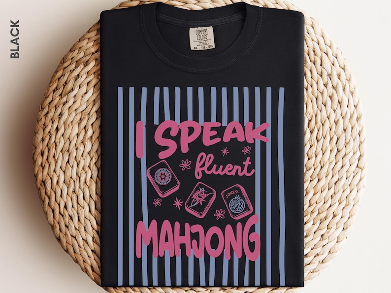 May include: A black t-shirt featuring a graphic with vertical light blue stripes and the pink text "I SPEAK fluent MAHJONG". The design includes illustrations of three mahjong tiles, including one labeled "JOKER", and small pink flower accents.