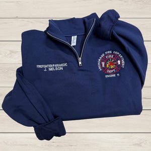May include: Navy blue fleece pullover with a zipper. The pullover has an embroidered patch on the front that says "Anchorage Fire Department" with a fire engine and the words "Fire Dept. Engine 11". The pullover also has the text "Firefighter/Paramedic J. Nelson" embroidered on the front.