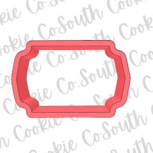May include: A red cookie cutter in the shape of a decorative label or tag. The cutter has a rectangular center with curved edges and a scalloped border. The background has a watermark of the words "Cookie Co. South".