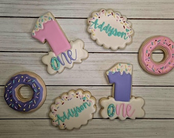 May include: Decorated sugar cookies in the shape of a number one, donuts, and a scalloped oval. The cookies are decorated with pastel colors, sprinkles, and the name "Addyson". The word "one" is also written on some of the cookies.