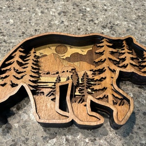 May include: A wooden bear silhouette wall art with a layered design. The bear's silhouette is cut out to reveal a scene of a mountain range, trees, and a moon in the sky.