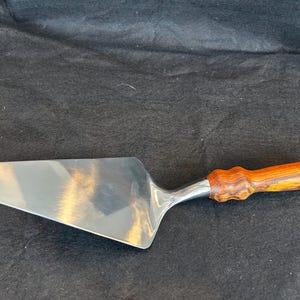 May include: A stainless steel pie server with a wooden handle. The server has a wide, triangular blade and a handle made of light and dark brown wood. The handle has a carved, textured design.