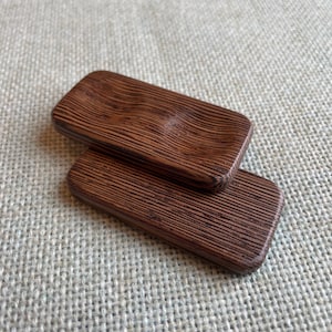 May include: Two rectangular wooden soap dishes with a dark brown, textured finish. One dish is stacked on top of the other. The top dish has a slight indentation. The wood grain is visible.