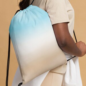 以下が含まれることがあります： グラデーションデザインの巾着バックパック。上部は水色で、中央は白、下部はベージュに変化します。黒い紐がデザインを完成させています。バッグは人が持っています。