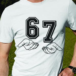 May include: White t-shirt featuring the number "67" in a bold, black, collegiate-style font. Below the numbers are two open hands, also in black, as if holding the numbers. The shirt is plain white.