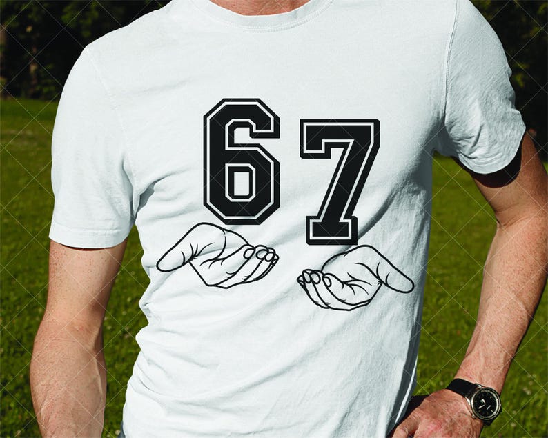 May include: White t-shirt featuring the number "67" in a bold, black, collegiate-style font. Below the numbers are two open hands, also in black, as if holding the numbers. The shirt is plain white.