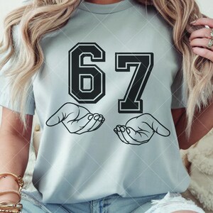 May include: Light blue t-shirt featuring the number 67 in a bold, black, collegiate-style font. Below the numbers are two open hands in a line drawing. The shirt is a casual, everyday wear item.