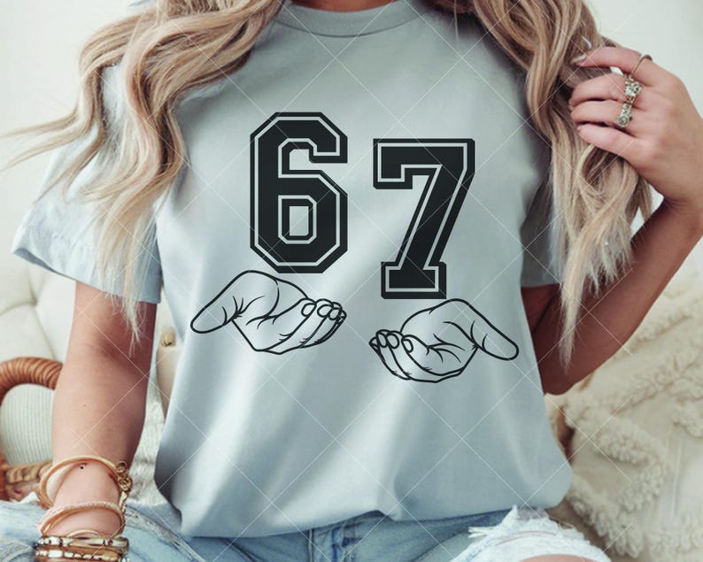 May include: Light blue t-shirt featuring the number 67 in a bold, black, collegiate-style font. Below the numbers are two open hands in a line drawing. The shirt is a casual, everyday wear item.