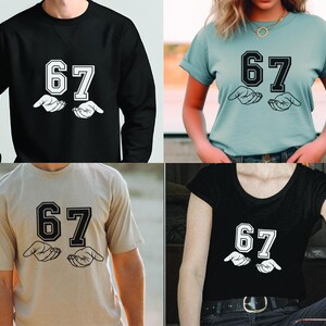 May include: Four different t-shirts and a sweatshirt are displayed. Each garment features the number "67" in a bold, outlined font above a graphic of two open hands. The shirts are in black, light blue, tan, and black.