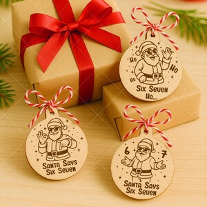 以下が含まれることがあります： サンタクロースのイラストと「Santa Says Six Seven」の文字が入った木製のクリスマスオーナメント。丸いオーナメントは赤と白の紐で結ばれています。背景には赤いリボンが付いたギフトボックスがあります。