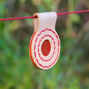 Puede incluir: Colgante de cuero hecho a mano con un diseño cosido en rojo y una abertura ovalada central. El colgante está suspendido por un cordón rojo sobre un fondo verde borroso. El cuero es de color marrón natural con detalles en rojo.