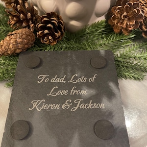 May include: A dark gray slate coaster with the inscription "To dad, Lots of Love from Kieron & Jackson" in elegant script. The coaster has four small, round, black pads on the bottom. The coaster is surrounded by pine cones and greenery.