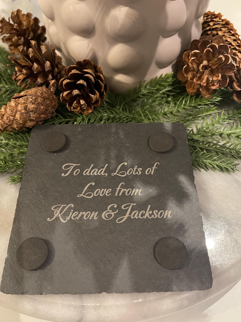 May include: A dark gray slate coaster with the inscription "To dad, Lots of Love from Kieron & Jackson" in elegant script. The coaster has four small, round, black pads on the bottom. The coaster is surrounded by pine cones and greenery.