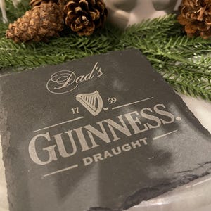 May include: A square, dark gray slate coaster with the Guinness logo and the words "Dad's" and "Draught" etched in white. The coaster is surrounded by pine cones and greenery.