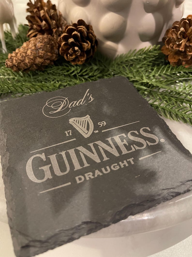 May include: A square, dark gray slate coaster with the Guinness logo and the words "Dad's" and "Draught" etched in white. The coaster is surrounded by pine cones and greenery.
