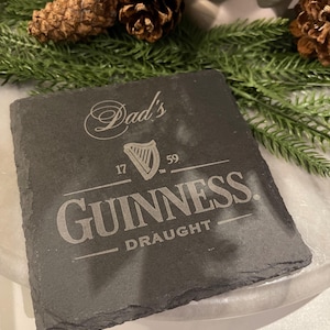 May include: A square, dark gray slate coaster with the Guinness logo and the word "Draught" engraved. The text "Dad's" is above the logo. The coaster is surrounded by pine needles and pine cones.