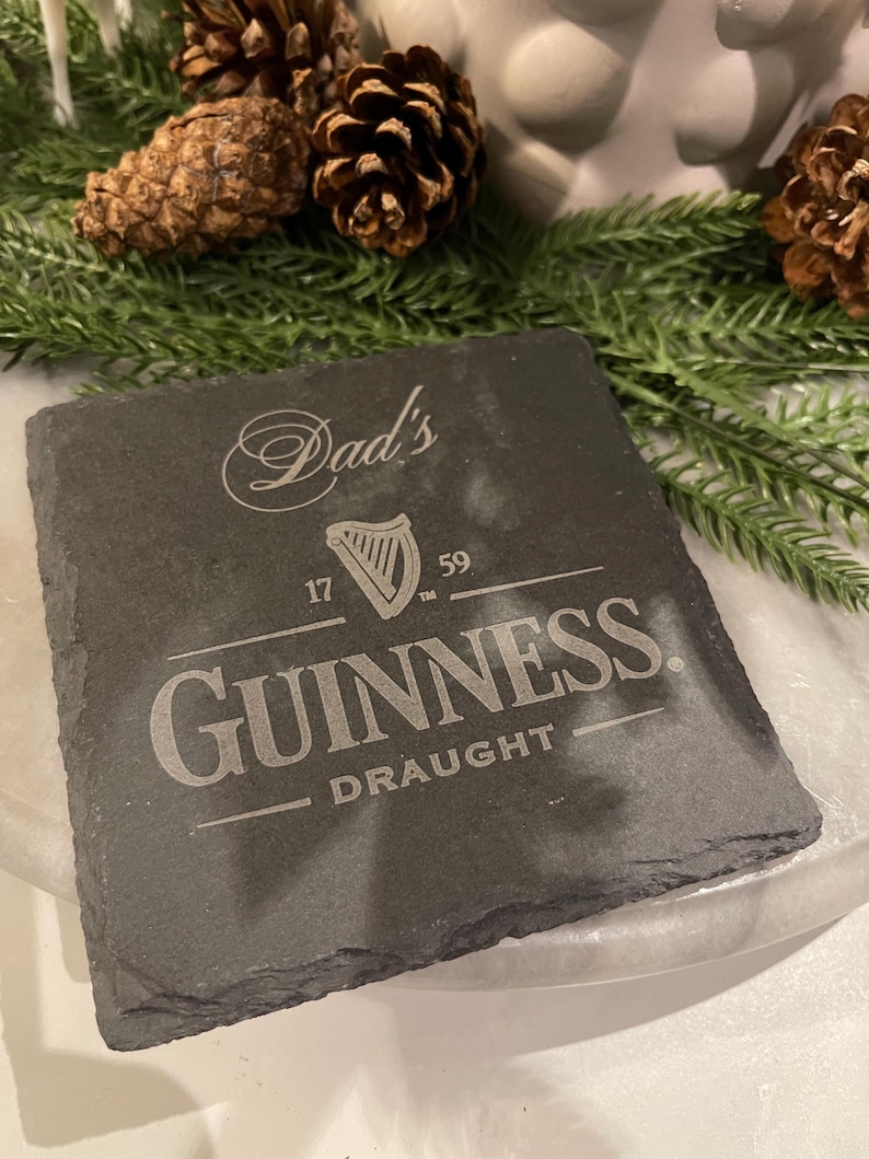 May include: A square, dark gray slate coaster with the Guinness logo and the word "Draught" engraved. The text "Dad's" is above the logo. The coaster is surrounded by pine needles and pine cones.