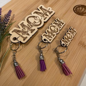 May include: Wooden keyrings with the word "MOM" and "I love you" engraved. Each keyring has a silver-coloured ring and a tassel in shades of purple. The keyrings are displayed on a wooden chopping board.