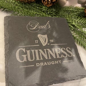 May include: A square, dark gray slate coaster with the Guinness logo and the words "Dad's" and "Draught" etched into the surface. The coaster also features a harp symbol and the numbers "17" and "59".