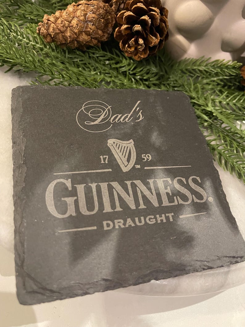 May include: A square, dark gray slate coaster with the Guinness logo and the words "Dad's" and "Draught" etched into the surface. The coaster also features a harp symbol and the numbers "17" and "59".