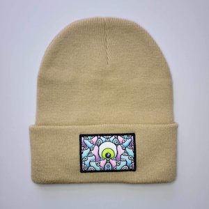May include: Beige beanie with a colorful eye patch featuring a green iris and pink and blue swirls.