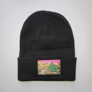 Puede incluir: Gorro negro con un parche colorido que representa un cactus y un paisaje desértico. El parche tiene un cielo rosa y un desierto marrón con un camino que lleva al cactus.