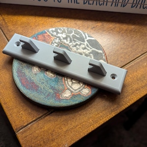 May include: A grey wall-mounted key holder with three hooks. The key holder is mounted on a wooden surface.