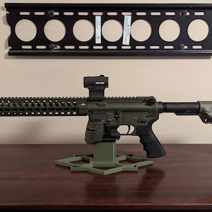 May include: An olive green rifle with a black handle and a red dot sight is displayed on a hexagonal stand. The rifle is positioned on a dark brown wooden surface, with a black wall mount and a light switch in the background.
