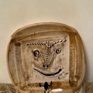 May include: A ceramic plate with a face design. The plate features a square face with a smiling mouth, eyes, and eyebrows. The plate is painted in shades of brown and beige, with black accents. The plate is a unique piece of art.
