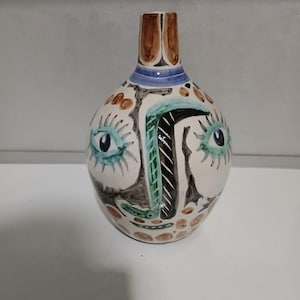Peut inclure: Un vase en céramique avec un motif de visage peint. Le vase présente une base blanche avec des accents marron, noirs, bleus et verts. Le motif du visage comprend deux yeux bleus, un nez et une bouche. Le haut du vase est marron.