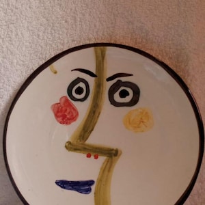 Puede incluir: Un plato de cerámica blanco con un borde marrón presenta un diseño de rostro abstracto pintado a mano. El rostro tiene ojos negros, una línea verde, labios rojos y acentos amarillos y rojos.