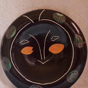 Peut inclure: Une assiette en céramique noire avec un motif de visage blanc. Le visage a deux yeux orange, un nez blanc et une bouche blanche. L'assiette est décorée de points verts et blancs autour du bord.
