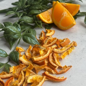 May include: Dried orange peels, arranged on a white surface, with fresh orange slices and green leaves in the background.