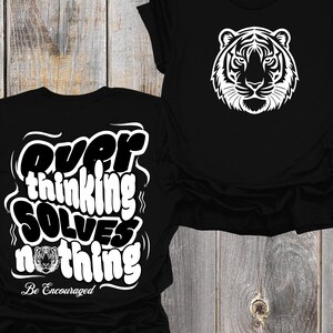 Puede incluir: Dos camisetas negras. Una presenta un gráfico de cara de tigre blanco. La otra tiene el texto "Overthinking Solves Nothing" en blanco, con un gráfico de tigre y las palabras "Be Encouraged".