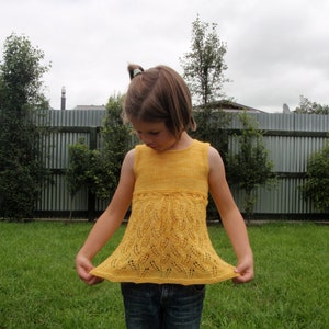 Puede incluir: Una blusa amarilla sin mangas de punto con un patrón de hojas detallado en la mitad inferior. La blusa tiene una forma ligeramente acampanada y un detalle de cordón en la cintura. La niña lleva vaqueros azules.
