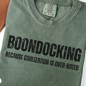 May include: A moss green t-shirt with the words "BOONDOCKING" and "BECAUSE CIVILIZATION IS OVER-RATED" printed in black. The shirt is made of a soft material and is displayed with a brown hat and dried plant stems.