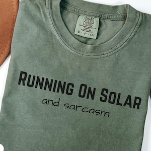 May include: Moss green t-shirt with the text "RUNNING ON SOLAR and sarcasm" in black. The shirt is a Comfort Colors brand, and the tag is visible. A brown hat and dried plant are in the background.