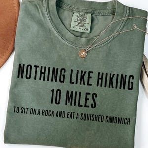 May include: A sage green Comfort Colors t-shirt with black text that reads 'NOTHING LIKE HIKING 10 MILES TO SIT ON A ROCK AND EAT A SQUISHED SANDWICH'. A gold necklace with a circular pendant rests on the shirt. A brown hat and dried leaves are also visible.