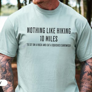 May include: A light green t-shirt with the text "NOTHING LIKE HIKING 10 MILES TO SIT ON A ROCK AND EAT A SQUISHED SANDWICH" in black. The shirt is a crew neck style and is being worn by a person.