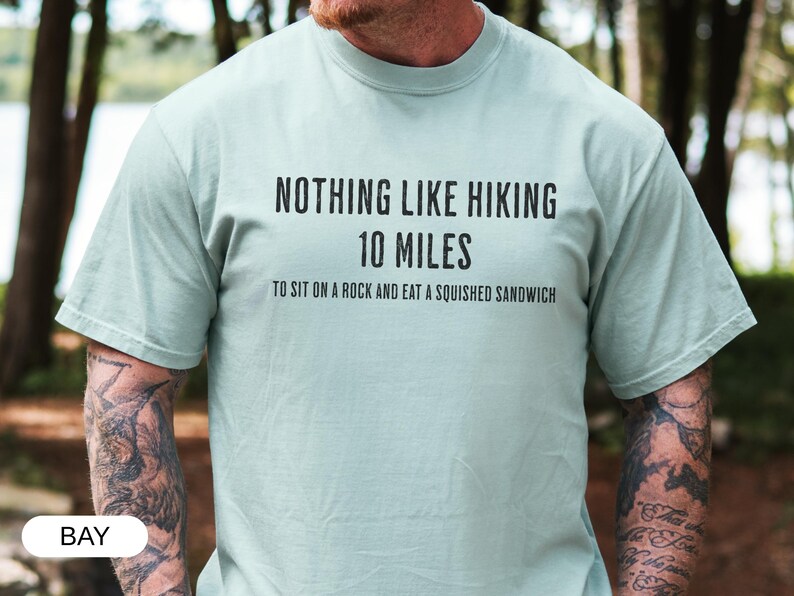 May include: A light green t-shirt with the text "NOTHING LIKE HIKING 10 MILES TO SIT ON A ROCK AND EAT A SQUISHED SANDWICH" in black. The shirt is a crew neck style and is being worn by a person.