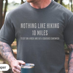 May include: Dark gray t-shirt with the text "NOTHING LIKE HIKING 10 MILES TO SIT ON A ROCK AND EAT A SQUISHED SANDWICH" in white. The shirt is a casual style, suitable for outdoor activities. The color is called "Pepper".