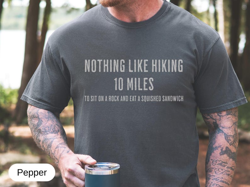 May include: Dark gray t-shirt with the text "NOTHING LIKE HIKING 10 MILES TO SIT ON A ROCK AND EAT A SQUISHED SANDWICH" in white. The shirt is a casual style, suitable for outdoor activities. The color is called "Pepper".
