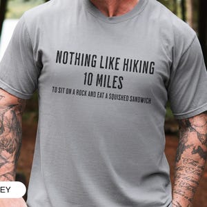 May include: Grey t-shirt with the text "NOTHING LIKE HIKING 10 MILES TO SIT ON A ROCK AND EAT A SQUISHED SANDWICH" in black. The shirt is short-sleeved and made of a soft material. The word "GREY" is at the bottom.