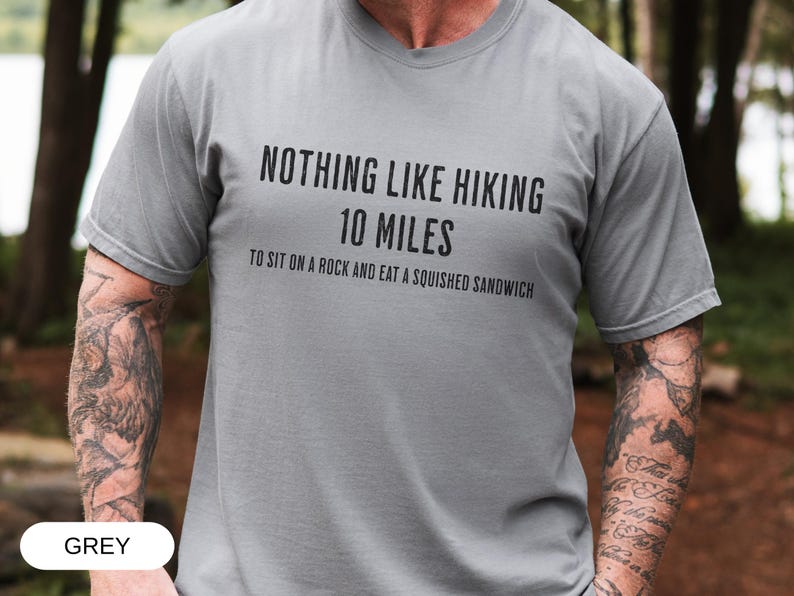 May include: Grey t-shirt with the text "NOTHING LIKE HIKING 10 MILES TO SIT ON A ROCK AND EAT A SQUISHED SANDWICH" in black. The shirt is short-sleeved and made of a soft material. The word "GREY" is at the bottom.