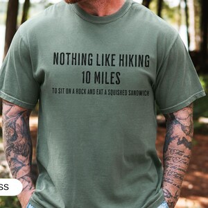 May include: Moss green t-shirt with the text "NOTHING LIKE HIKING 10 MILES TO SIT ON A ROCK AND EAT A SQUISHED SANDWICH" in black. The shirt is a casual style, suitable for outdoor activities.